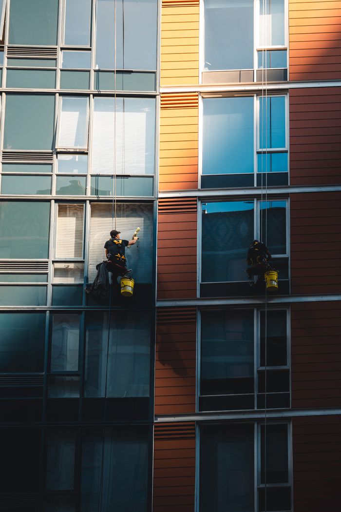 services-03 Two workers hanging to clean windows on a modern skyscraper facade.