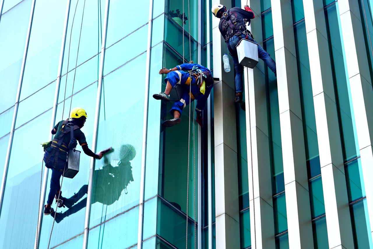 hero-img-02 Three window cleaners suspended on ropes clean a modern glass building facade during the day.